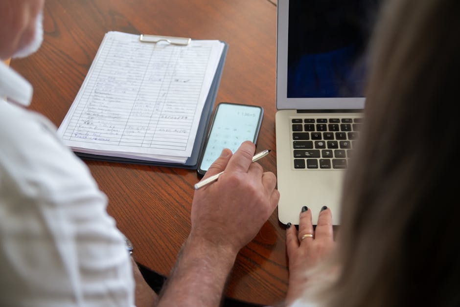 Two adults working at a desk with a laptop, smartphone, and documents. Collaborative workspace scene.